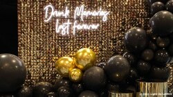 A decorative wall with a neon sign reading "Drunk Moments Last Forever," surrounded by black and gold balloons.