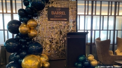 Decorative event stage with a backdrop of golden sequins, a sign reading "VIP Barrel Event," and clusters of blue and gold balloons. Two chairs are placed to the side.
