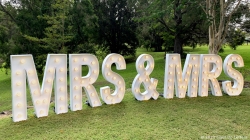 Large illuminated letters spelling "MRS & MRS" on a grassy area with trees in the background.