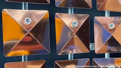 Close-up of shiny copper-colored square panels with raised centers and small round attachments, arranged in a grid against a dark background.
