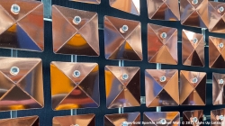 Close-up of a grid of copper-colored, square metal panels with a shiny, reflective surface, each featuring a central rivet or fastener.