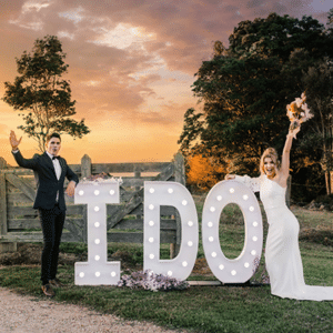 A couple celebrates their wedding beside large, illuminated "I DO" letters outdoors at sunset, with trees and a wooden fence in the background.