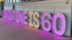 Illuminated sign displaying "CHRISTINE IS 60" with purple and white lights against a wall.