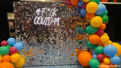 A sign with "#F*CK COVID-19" in illuminated text is surrounded by colorful balloons.