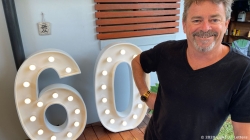 Man smiling next to large, illuminated "60" sign at an outdoor gathering.