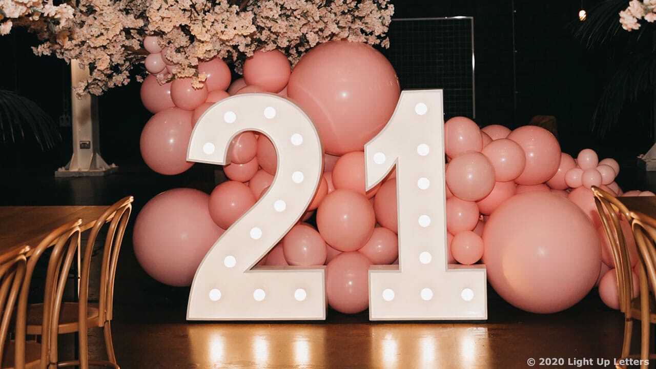 Large illuminated "21" sign stands in front of a pink balloon arrangement at an indoor event. Chairs and tables are set up nearby.
