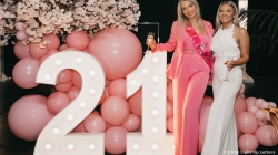 Two women stand near large lit "21" numbers, surrounded by pink balloons and flowers. One wears a pink sash and suit, the other in a white outfit, both holding drinks.